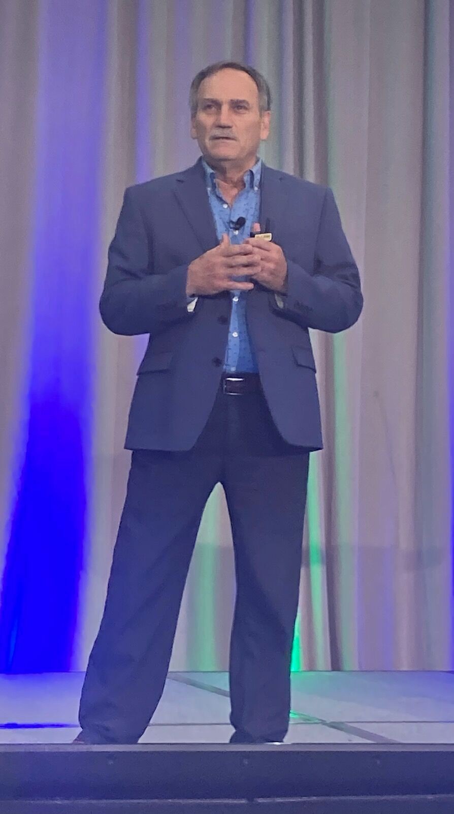 Man in a suit speaking on stage with blue and green lighting in the background.