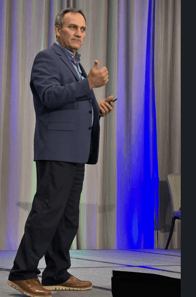 Person in a blue suit standing on stage, holding a microphone and giving a thumbs-up gesture.